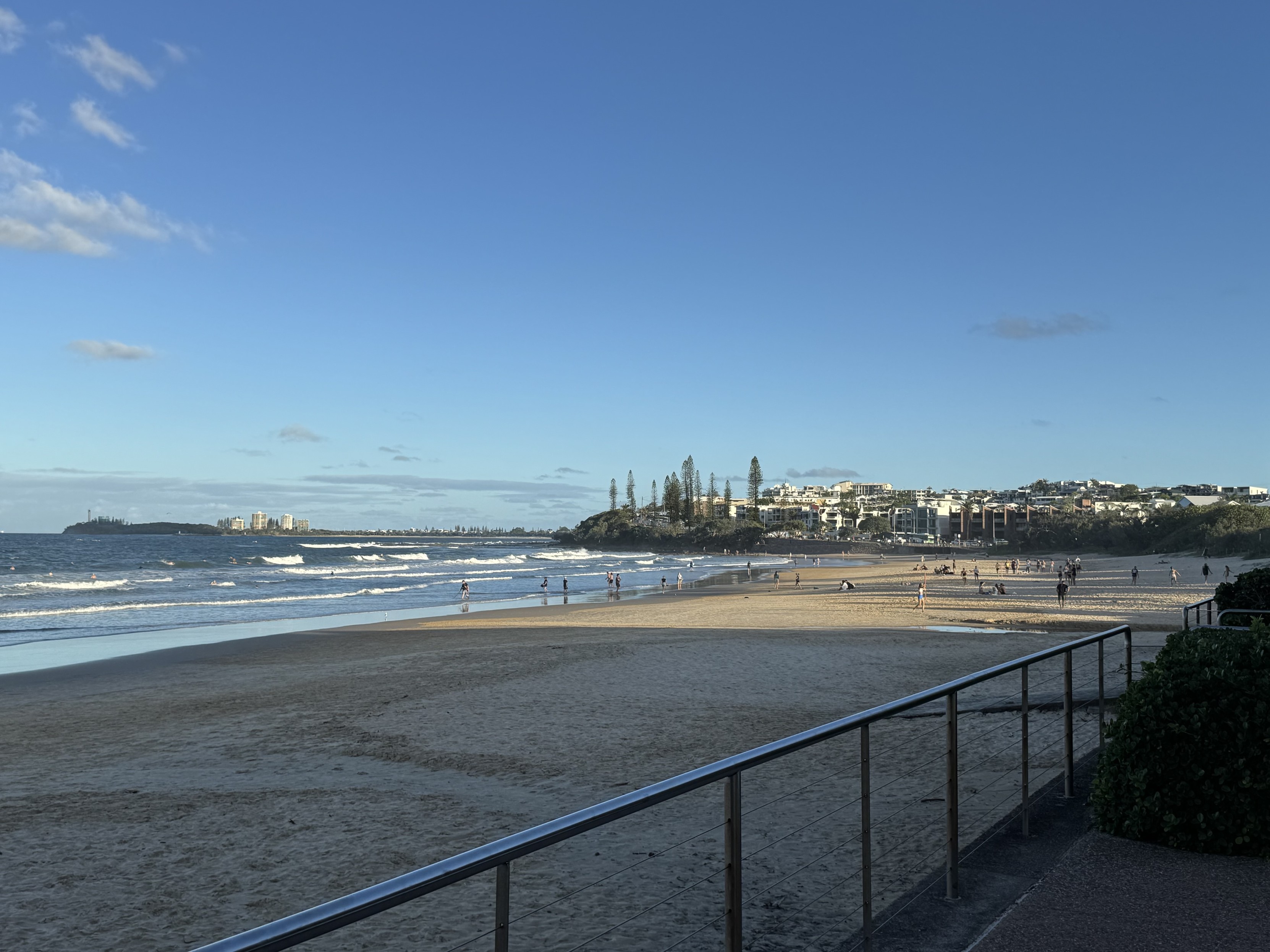 Looking at Alexandra Headland from a steel railing, with a long stretch of sandy beach in between. The tide is out and late afternoon shadows are cast onto the sand from buildings not in view. There are low rise buildings on the headland, dotted with tall Norfolk Pine trees. Very few clouds, most low on the horizon out to sea.